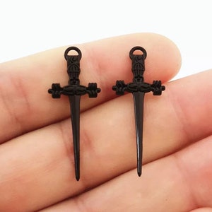 May include: Two black metal sword charms with intricate details. The swords have a pointed tip and a cross-shaped guard.