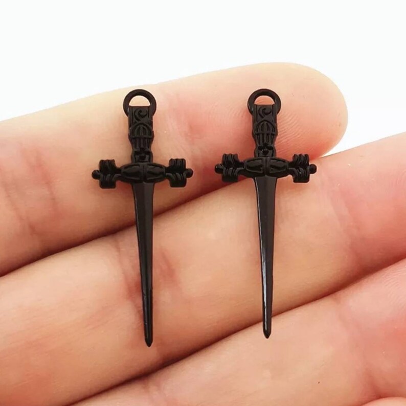 May include: Two black metal sword charms with intricate details. The swords have a pointed tip and a cross-shaped guard.
