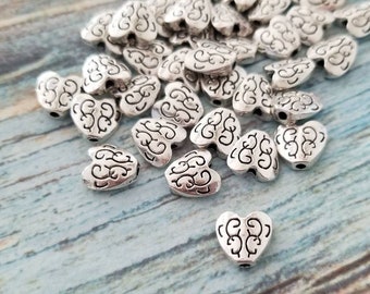May include: A pile of silver heart-shaped beads with a decorative swirl design. The beads have a hole in the top for stringing.