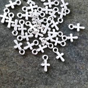 Op de afbeelding: Een stapel zilverkleurige metalen kruisbedels met een lus aan de bovenkant. De bedels liggen verspreid op een grijs, getextureerd oppervlak.