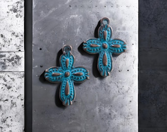May include: Two turquoise-colored cross pendants with a bronze-colored trim. The crosses have a detailed design with a loop at the top for hanging. The background is a textured, gray surface.
