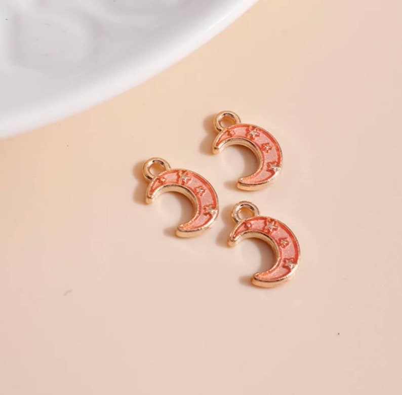 Op de afbeelding: Drie goudkleurige halve maan bedels met een roze emaille afwerking en ster details. Elke bedel heeft een kleine lus aan de bovenkant om aan sieraden te bevestigen. De bedels worden gepresenteerd op een lichtroze oppervlak.