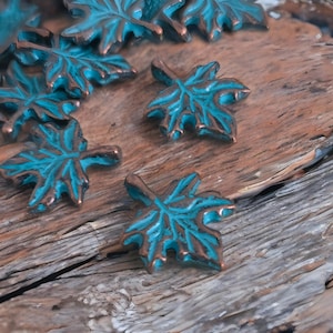 May include: Close-up of several teal and copper-colored maple leaf charms. The charms have a distressed finish, with teal highlighting the leaf veins and copper outlining the edges. The charms are scattered on a weathered wooden surface.