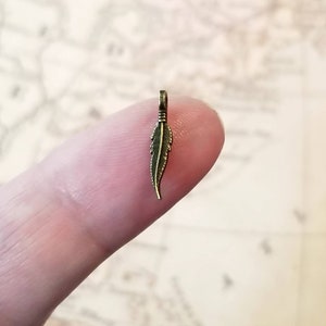 May include: A small, gold-toned feather charm pendant. The feather is detailed with lines to resemble a real feather.