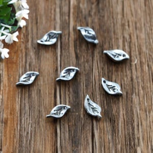 May include: A collection of small, silver-colored leaf charms scattered on a wooden surface. The charms have detailed veining and a slightly textured appearance. The background is a warm, brown wood grain.