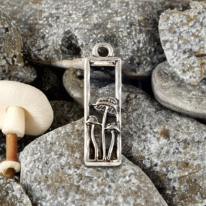 May include: Silver charm with a rectangular frame and a design of three mushrooms inside. The charm has a loop at the top for attaching to a necklace or bracelet.