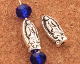 May include: Two silver-toned Virgen de Guadalupe charms with a blue crystal bead on each side. The charms are oval-shaped and feature a detailed image of the Virgin Mary.