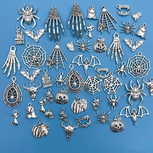 May include: A collection of silver-toned metal charms featuring various Halloween-themed designs including spiders, spiderwebs, skeleton hands, bats, pumpkins, ghosts, skulls, cats, and vampire teeth, all arranged on a solid light blue background.