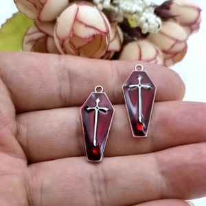 Op de afbeelding: Twee zilverkleurige doodskist-charms met rood emaille en een zilveren kruisontwerp.