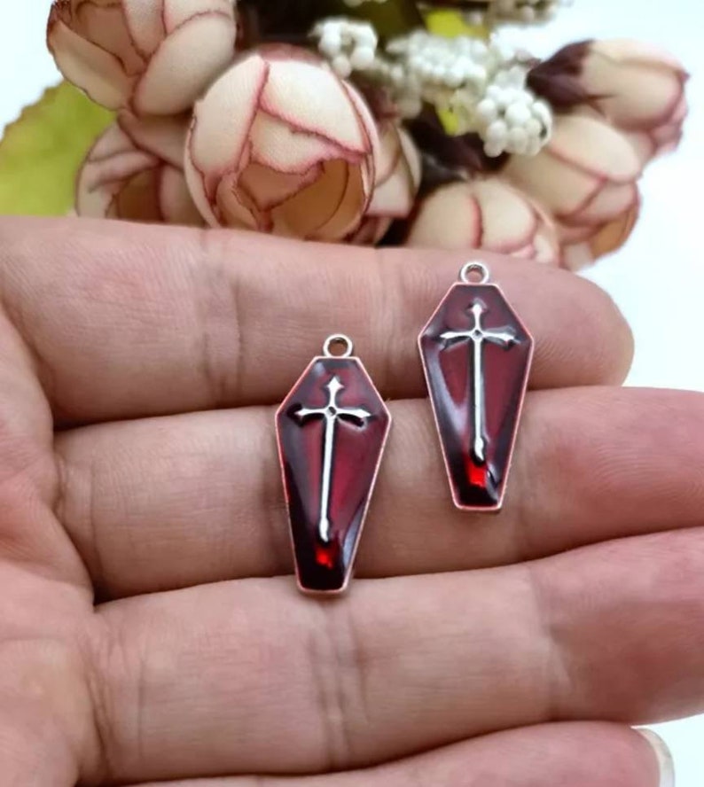 Op de afbeelding: Twee zilverkleurige doodskist-charms met rood emaille en een zilveren kruisontwerp.
