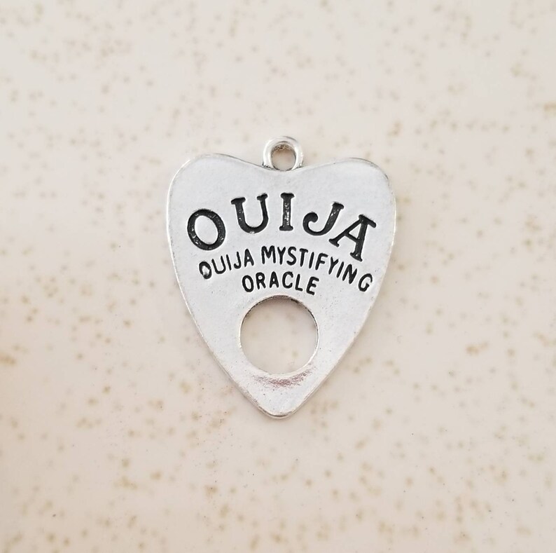 May include: Silver charm in the shape of a heart with the word "OUIJA" written on it. The charm also says "OUIJA MYSTIFYING ORACLE".