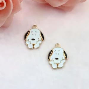 Op de afbeelding: Twee Snoopy-vormige bedels. Elke bedel heeft een witte body, zwarte oren en een goudkleurige omtrek. De bedels hebben een kleine lus aan de bovenkant om aan sieraden of andere knutselwerken te bevestigen. De achtergrond is zachtroze.