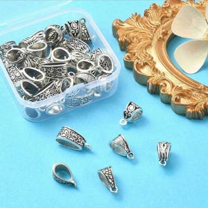 May include: A collection of silver-colored bail connectors with intricate, embossed designs. Some are in a clear plastic container, while others are scattered on a light blue surface. These jewelry findings have small loops for attaching to pendants or other components.