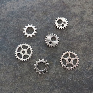 May include: Six silver metal gears of varying sizes and designs. The gears are arranged in a semi-circle on a gray background.