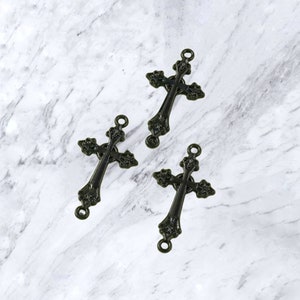 May include: Three bronze-colored metal crosses with ornate detailing. Each cross has a loop at the top and bottom for attaching to other objects. The crosses are arranged on a white and gray marble-patterned surface.