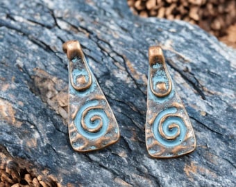 Puede incluir: Dos colgantes de color bronce con un diseño de espiral turquesa. Los colgantes tienen forma triangular con un lazo en la parte superior para colgar. Los colgantes tienen un aspecto envejecido y desgastado.