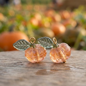 Op de afbeelding: Twee pompoenvormige bedels gemaakt van doorschijnend oranje glas met gouden glitters aan de binnenkant. Elke bedel heeft een geribbelde textuur en een klein, donkergroen glazen blaadje bevestigd aan een goudkleurige metalen ring.