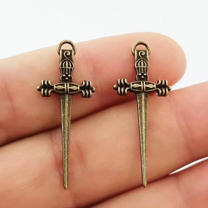 May include: Two antique gold-toned sword charms with intricate details. The swords have a pointed tip and a decorative cross guard.