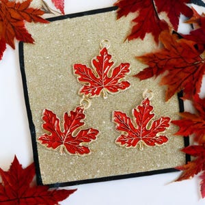 May include: Three red maple leaf charms with gold-colored trim and a small loop for hanging. The charms are set on a textured, light brown surface, surrounded by red autumn leaves.