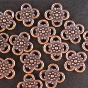 May include: A collection of 14 copper-colored metal charms in the shape of a four-leaf clover. Each charm has a decorative design in the center.