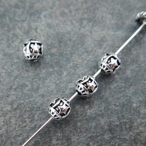 May include: A set of round, silver-toned metal beads featuring an intricate cut-out design of stars and crescent moons. Four of these spherical beads are threaded onto a thin metal wire, while one additional matching bead sits separately nearby.
