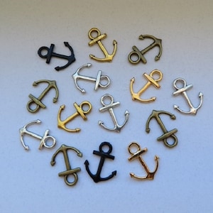 K&ouml;nnte beinhalten: Eine Sammlung von 13 silbernen, goldenen und schwarzen Anker-Anh&auml;ngern. Die Anh&auml;nger sind in einem Kreis auf wei&szlig;em Hintergrund angeordnet.