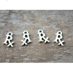 May include: Five silver-toned metal charms shaped like the medical symbol "Rx". The charms are arranged in a row on a wooden surface.