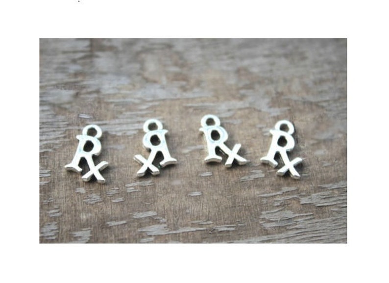 May include: Five silver-toned metal charms shaped like the medical symbol "Rx". The charms are arranged in a row on a wooden surface.