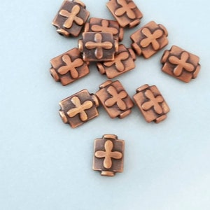 May include: A group of copper-colored metal beads with a cross design. The beads are scattered on a light blue surface.