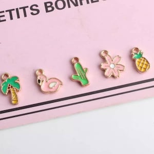 May include: Five gold-colored metal charms with enamel paint. The charms are shaped like a palm tree, flamingo, cactus, flower, and pineapple. The charms are on a pink surface.