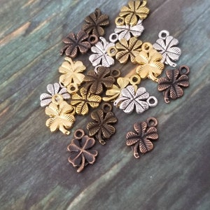 May include: A collection of 16 gold, silver, and bronze four-leaf clover charms with a loop at the top. The charms are scattered on a wooden surface.