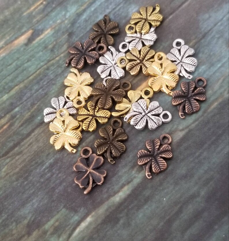 May include: A collection of 16 gold, silver, and bronze four-leaf clover charms with a loop at the top. The charms are scattered on a wooden surface.