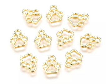 May include: Ten gold-colored metal paw print charms with a loop at the top. The charms are all the same size and shape, and they are arranged in a random pattern on a white background.