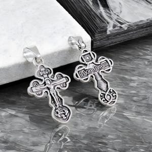 May include: Two silver-colored Orthodox crosses with intricate detailing. One cross features a depiction of Jesus Christ, while the other has Cyrillic text. Both have a loop for a chain.