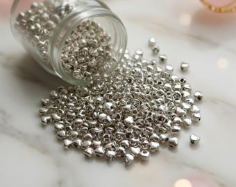Könnte beinhalten: Ein durchsichtiges Glasgefäß, das zur Seite geneigt ist und eine große Menge kleiner, metallisch silberner herzförmiger Perlen ausschüttet. Die Perlen haben eine glatte, reflektierende Oberfläche und ein Loch in der Mitte.