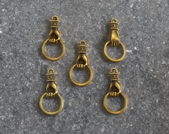 May include: Five antique gold-colored charms. Each charm features a hand holding a ring, with a decorative top. The charms are arranged on a neutral surface, showcasing their intricate details and vintage aesthetic. Ideal for jewelry making.
