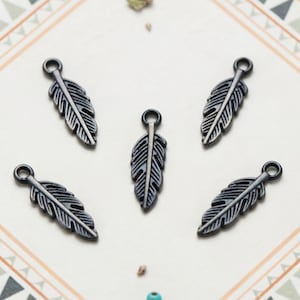 May include: Five identical dark gunmetal gray feather charms arranged in a circular pattern. Each charm features a detailed central quill and textured barbs with a small circular loop at the top for attachment, showcasing a metallic finish.