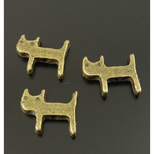 Puede incluir: Tres peque&ntilde;os amuletos de metal de color dorado antiguo con forma de gato. Los amuletos son simples y estilizados, con orejas redondeadas y un peque&ntilde;o c&iacute;rculo para cada ojo.