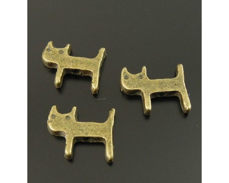 Puede incluir: Tres peque&ntilde;os amuletos de metal de color dorado antiguo con forma de gato. Los amuletos son simples y estilizados, con orejas redondeadas y un peque&ntilde;o c&iacute;rculo para cada ojo.