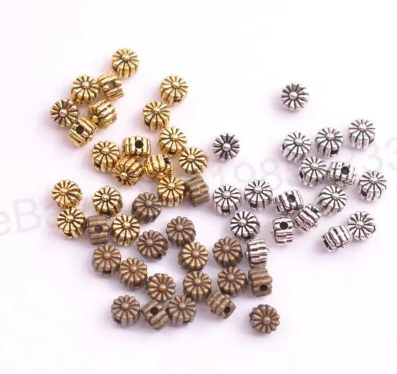 May include: A collection of small, round, flower-shaped metal beads in gold, silver, and bronze tones. Each bead features a radial, ribbed pattern with a central hole, arranged in a scattered cluster on a plain white background.