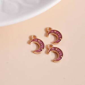Op de afbeelding: Drie goudkleurige halve maan bedels met een paarse emaille vulling en ster details. Elke bedel heeft een kleine lus aan de bovenkant om aan sieraden of andere knutselwerken te bevestigen. De bedels liggen op een lichtroze ondergrond.