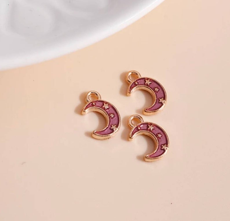 Op de afbeelding: Drie goudkleurige halve maan bedels met een paarse emaille vulling en ster details. Elke bedel heeft een kleine lus aan de bovenkant om aan sieraden of andere knutselwerken te bevestigen. De bedels liggen op een lichtroze ondergrond.