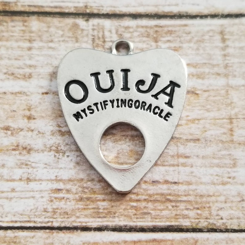 May include: A silver charm in the shape of a heart with the word "OUIJA" and the text "MYSTIFYING ORACLE" around the edge. The charm has a hole in the center.