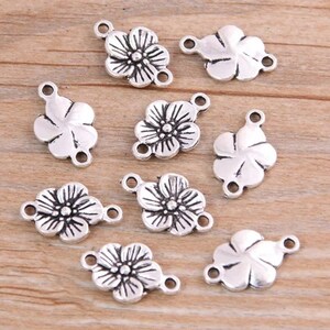 Puede incluir: Diez abalorios de flores plateados con dos anillos de salto cada uno. Los abalorios tienen forma de flor con cinco p&eacute;talos y un detalle central.