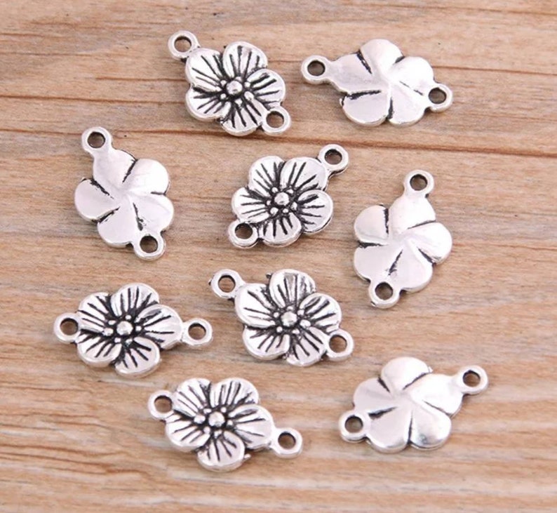 Puede incluir: Diez abalorios de flores plateados con dos anillos de salto cada uno. Los abalorios tienen forma de flor con cinco p&eacute;talos y un detalle central.
