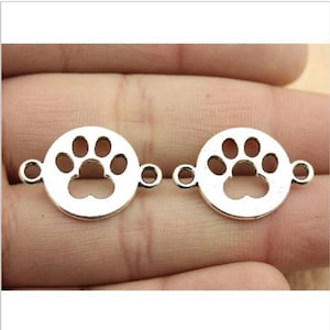 May include: Two silver-toned metal charms in the shape of a paw print. Each charm has a circular design with a paw print cut out in the center. The charms have two jump rings on each side for attaching to jewelry or other crafts.