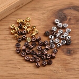 May include: A collection of small, round metal beads featuring a radial, flower-like pattern on the top and bottom surfaces. The beads are finished in three distinct metallic tones: polished gold, bright silver, and a dark, weathered copper.