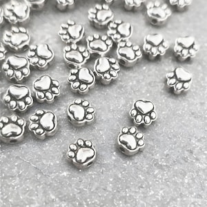 May include: A collection of small, silver-colored paw print beads. Each bead features a detailed paw print design, with rounded toe pads and a heart-shaped center. The beads are scattered across a light gray surface.