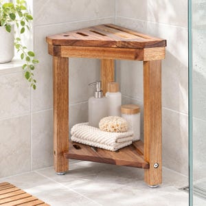 May include: A triangular wooden shower stool with a slatted top and a lower shelf. The shelf holds a white pump bottle, a white cylindrical bottle, a textured beige sponge, and a clear jar with a wooden lid, all resting on a folded white towel.