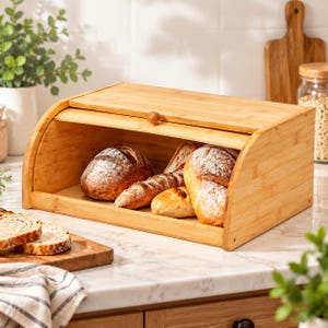 May include: A rectangular bamboo bread box with a roll-top lid and a small wooden knob. The interior contains several loaves of artisan bread, including a round sourdough loaf, a baguette, and a smaller roll, all with a golden-brown crust.